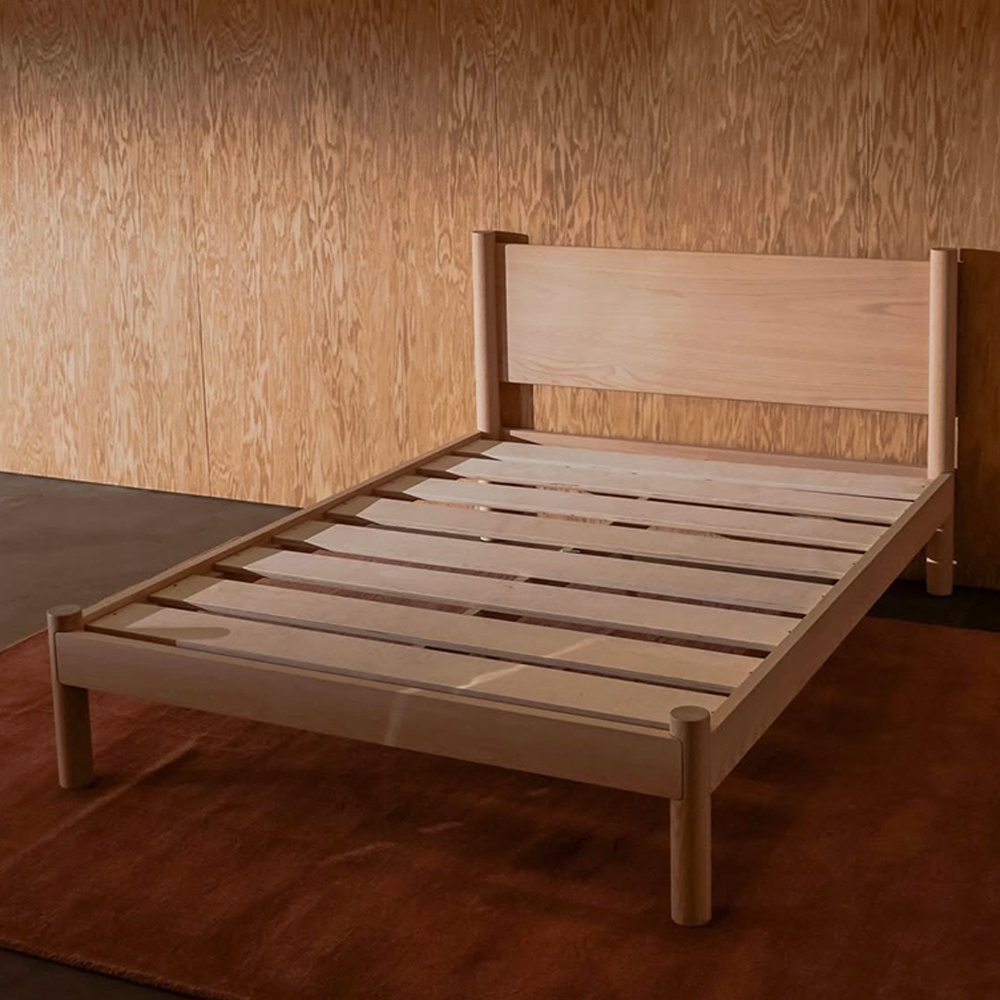 A light natural modern solid wood designer bed frame Keeps Architect bed with headboard integrated joinery, shown in a studio setting against a wood-paneled wall and a brown rug.
