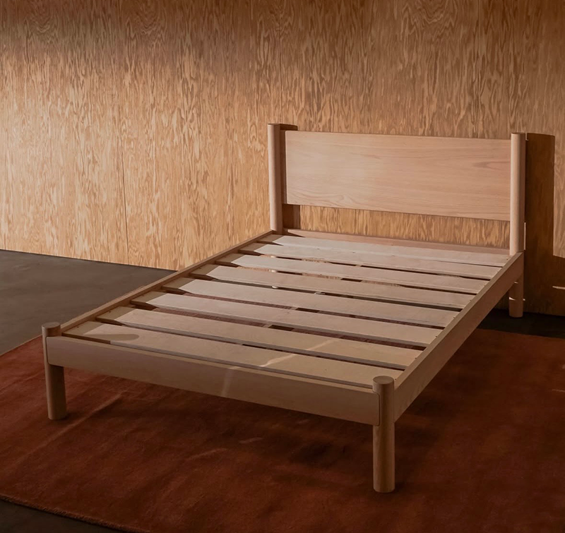 A light natural modern solid wood designer bed frame Keeps Architect bed with headboard integrated joinery, shown in a studio setting against a wood-paneled wall and a brown rug.