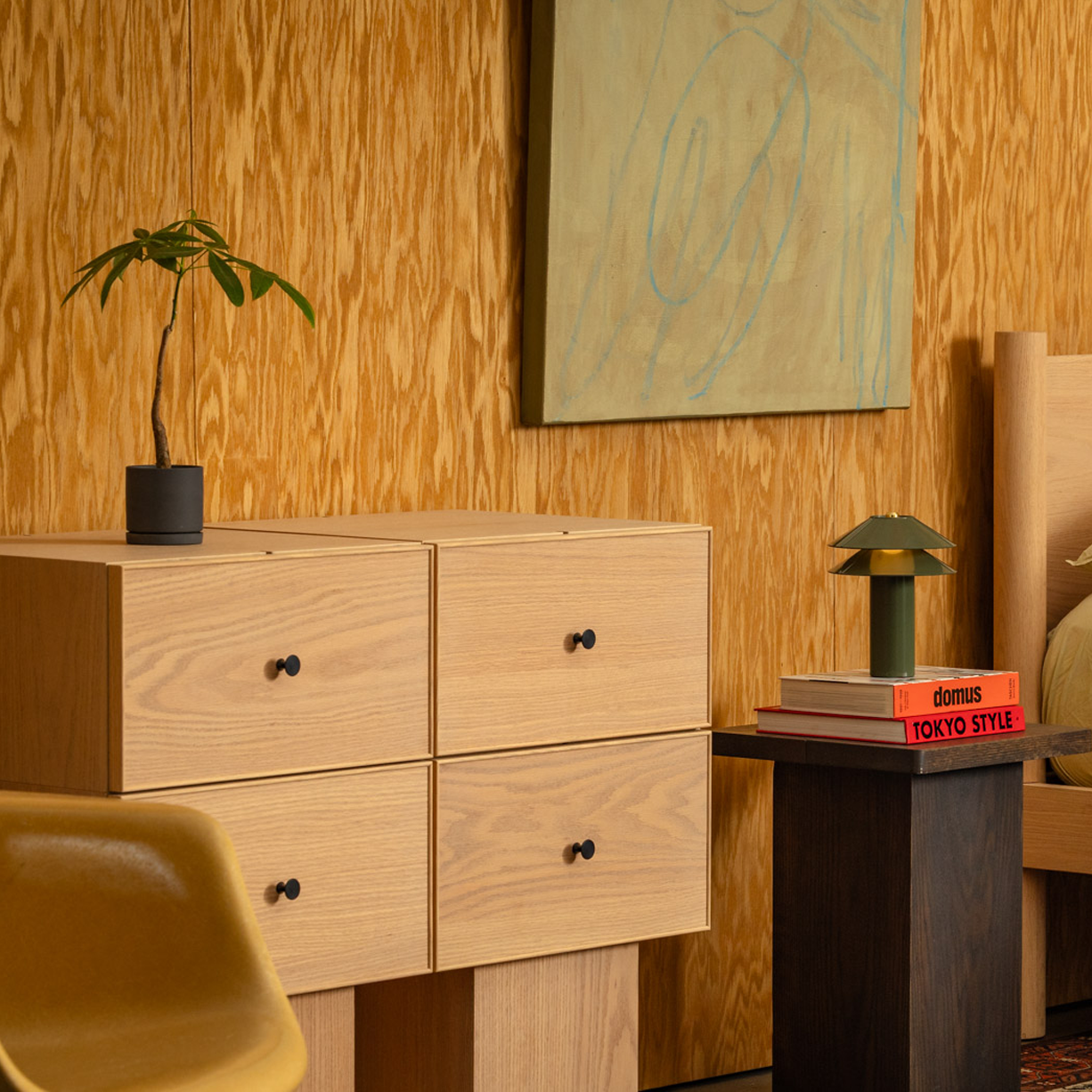 Keeps Anywhere oak four-drawer dresser modular storage in light natural finish with plant and red watering can, styled in a wooden mid-century bedroom with art, Keeps architect bed, and minimalist end table in smoke with Minale table light.