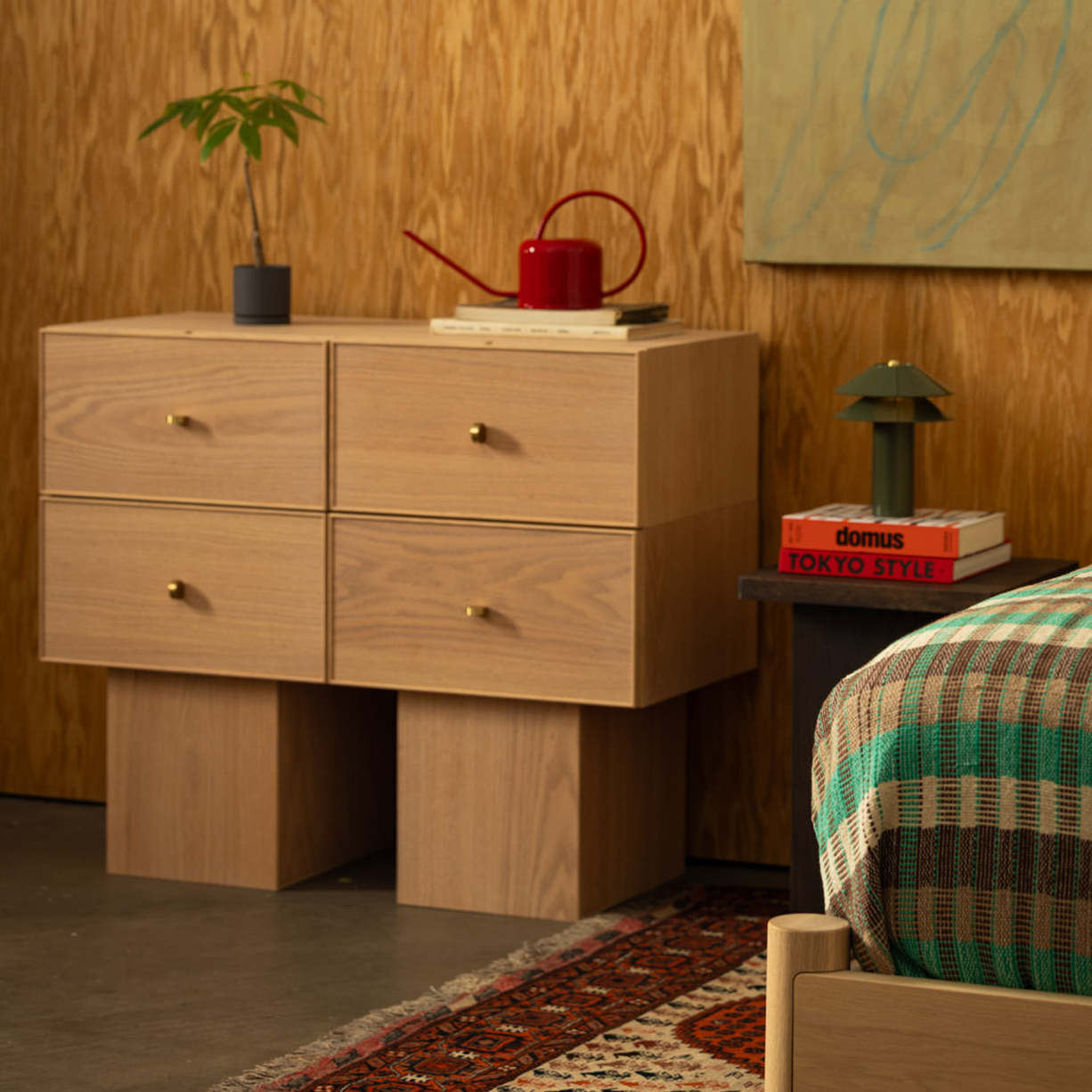 Keeps Anywhere oak four-drawer dresser modular storage in light natural finish with plant and red watering can, styled in a wooden mid-century bedroom with Keeps architect bed corner, and minimalist end table in smoke with Minale table light.