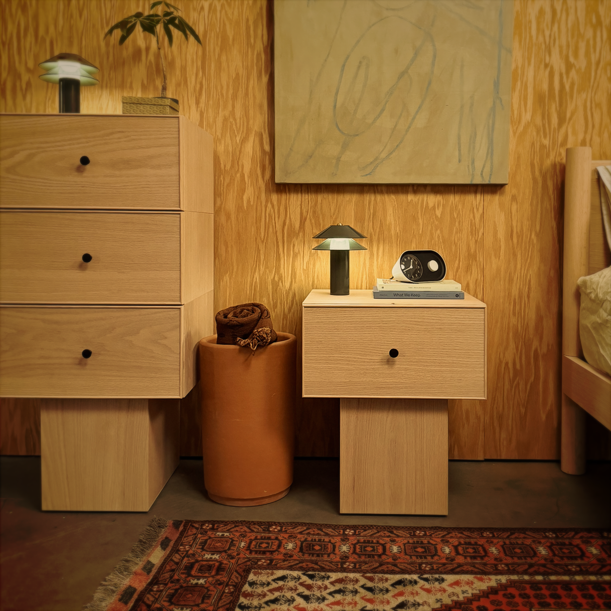 Keeps Anywhere wooden drawer nightstand and stackable three-drawer tall dresser in light natural finish, set against a wood-paneled wall with a painting, Minale lamps, and a vintage Afghan rug.