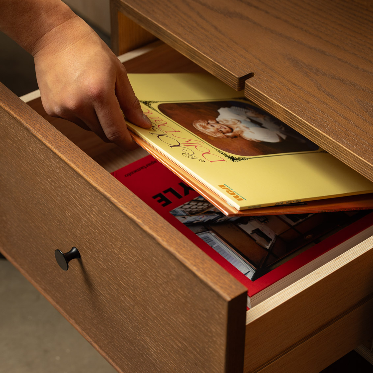 Close-up of Keeps Anywhere wooden drawer nightstand dresser with a black knob, partially open to reveal storage for vinyl records inside, on a matching wooden surface.