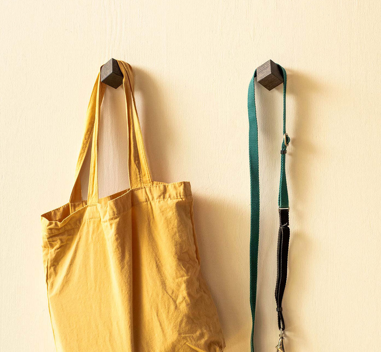 Yellow tote bag and green strap hanging on wooden hooks on a beige wall.