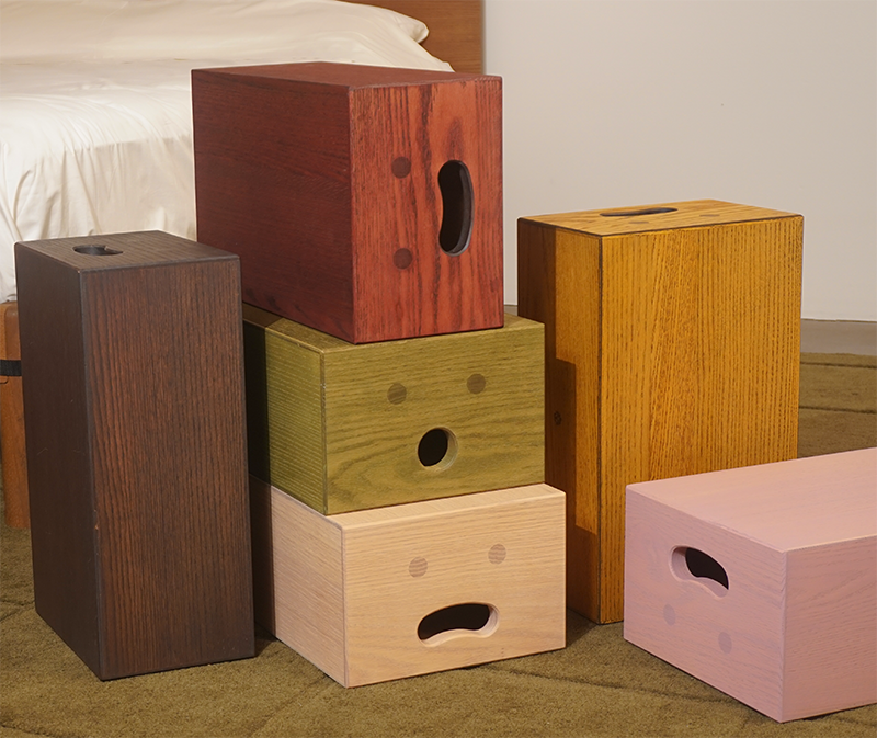 Six Keeps Off-Cuts Apple Boxes in different stained solid oak colors with natural linseed oil, stacked and arranged on a green carpeted floor in front of a bed.