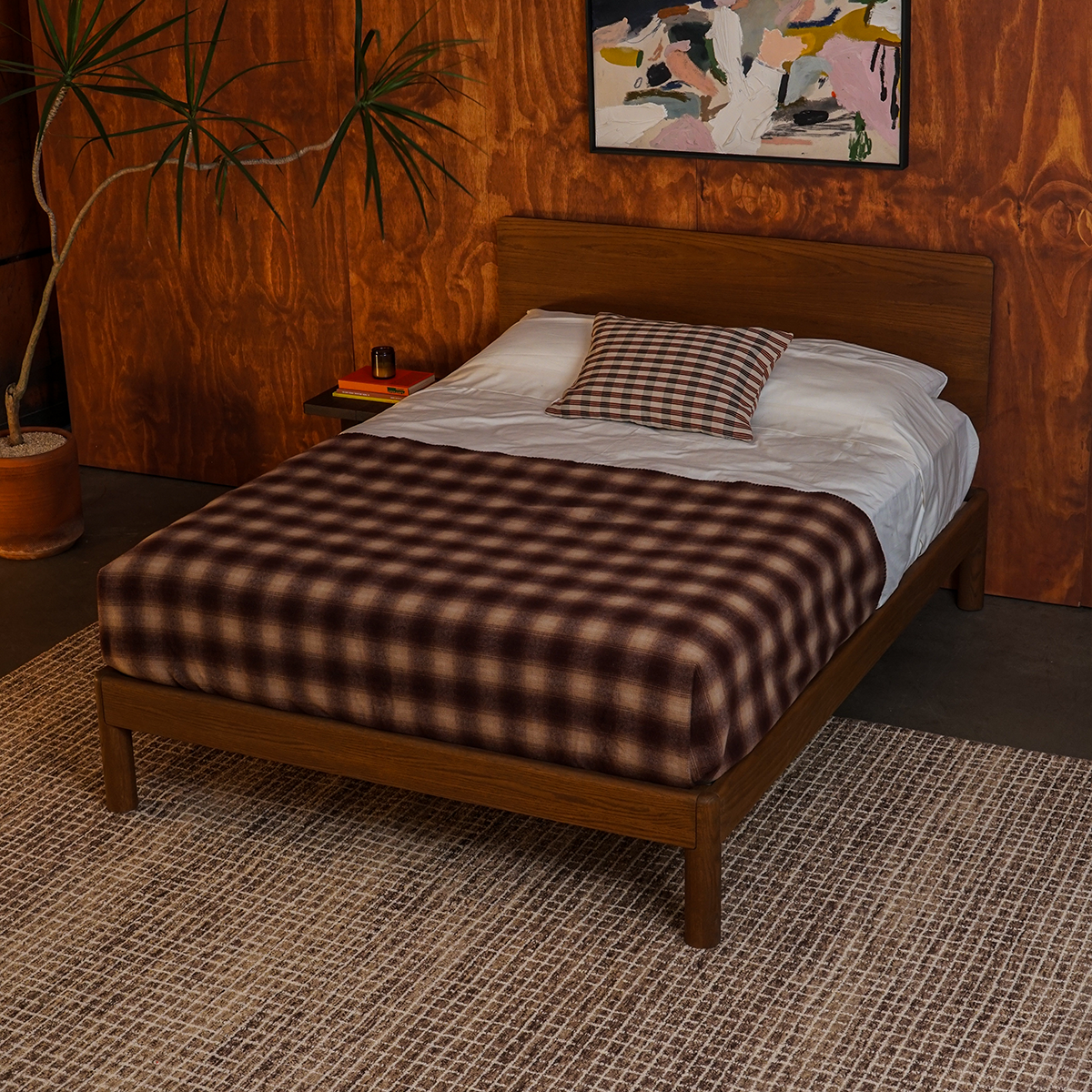 Keeps bolted oak bed frame in acorn brown finish with plaid blanket in a cozy room with wooden walls, spiky plant, abstract art, and checked rug.