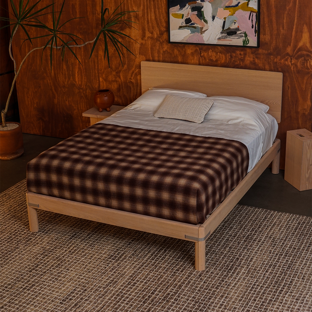 Keeps strap oak bed frame in natural finish with headboard and plaid blanket in a cozy room with wooden walls, spiky plant, two natural keeps nightstands, and checked rug.