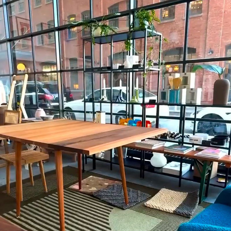 M Collection Home's modern showroom space in DUMBO with wooden table, chairs, and large windows showing an urban street view.